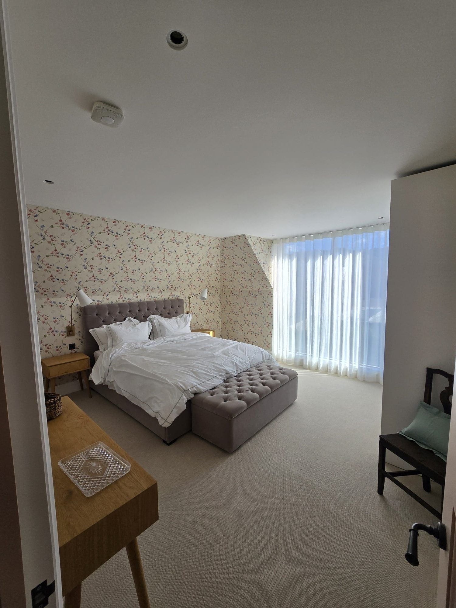 Primary bedroom finished with floral wallpaper, upholstered bed, and full-height sheer curtains.