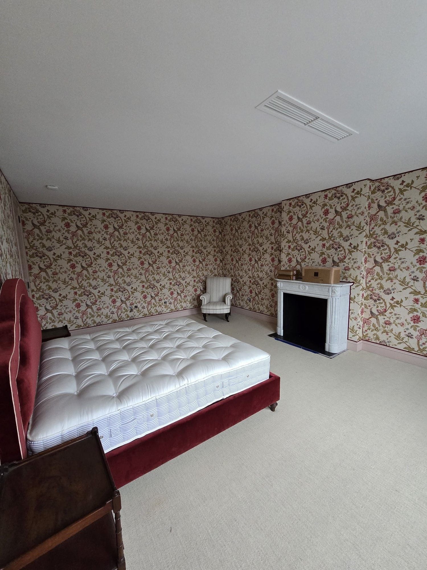 Bedroom fully wrapped in floral wallpaper with a fireplace, armchair, and light carpet.